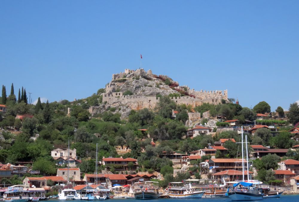 The castle, but mostly just its walls, on a peak above a small town on the waterfront, with boats.