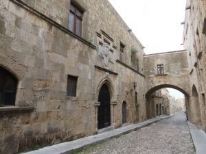 A cobbled street lined with stone 2-story buildings. Two of them are connected by a bridge over the street.