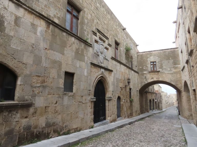 A cobbled street lined with stone 2-story buildings. Two of them are connected by a bridge over the street.