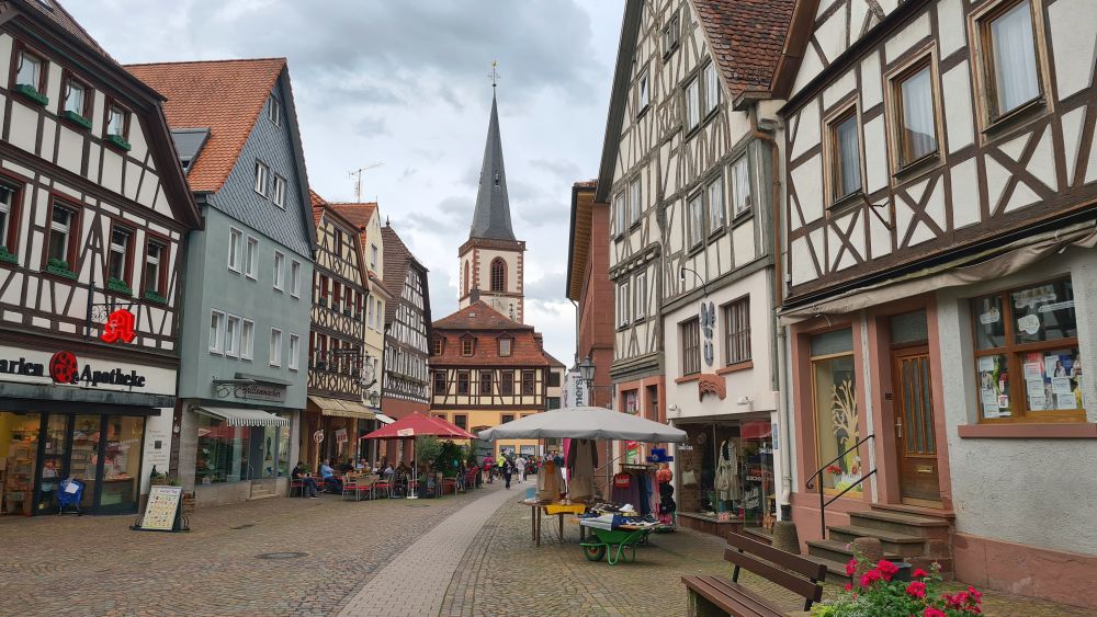 A pretty pedestrian street lined with half-timbered houses and shops, with outdoor cafe tables and umbrellas. A church tower in the background.