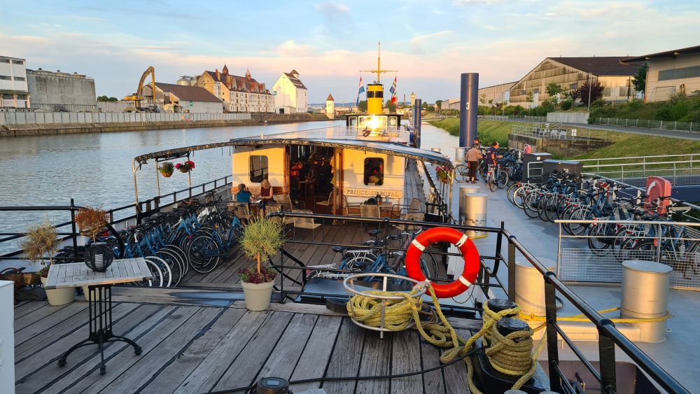 A view from the front of the boat toward the back: a wooden deck, a shaded area, and the indoor lounge beyond that. Bikes on the dock next to the ship.