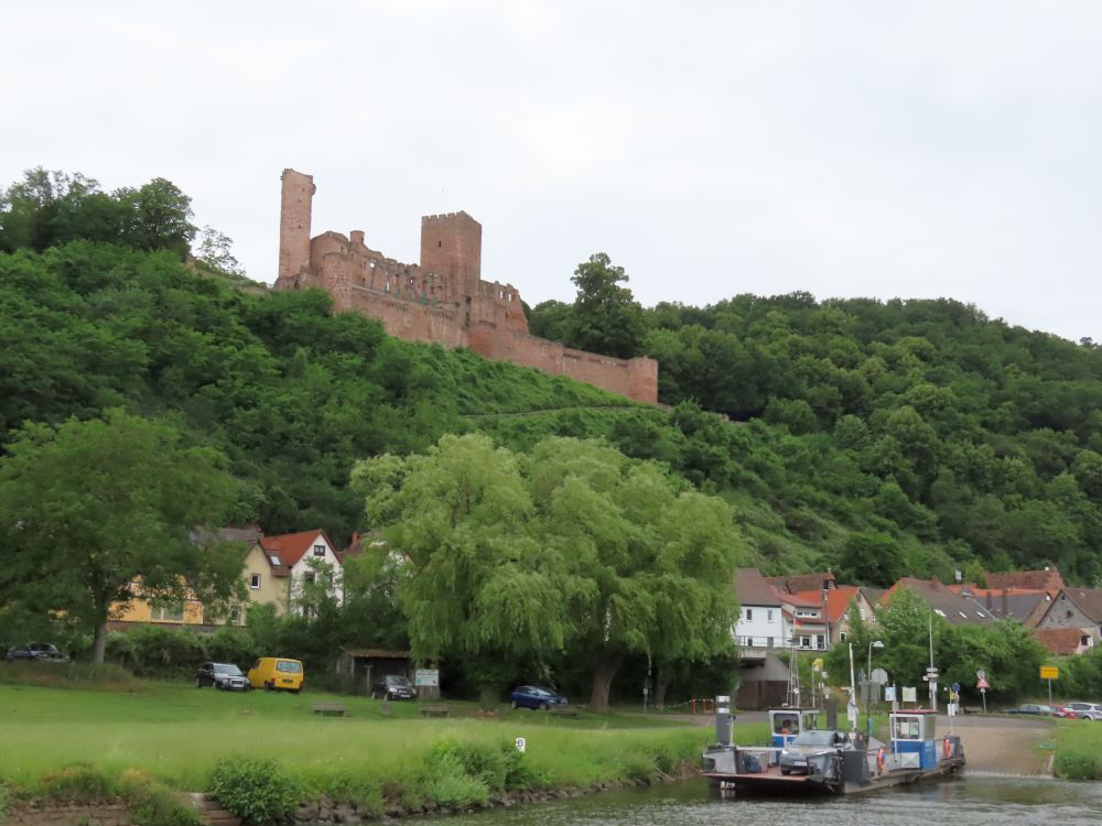 Houses at the base of a wooded hill, a large castle ruin on top of the hill, a very small car ferry parked on the edge of the river.
