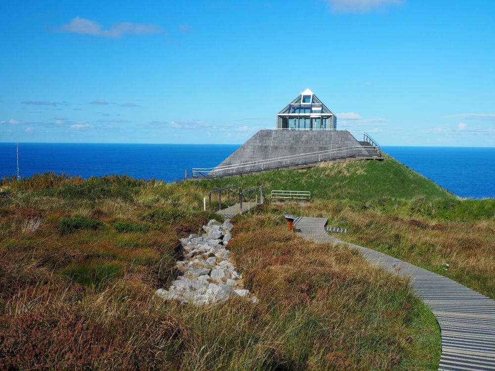 A triangular building with a glass-walled room at its top. In front, a boardwalk with the path of a stone wall visible next to it. Beyond the building, the sea.