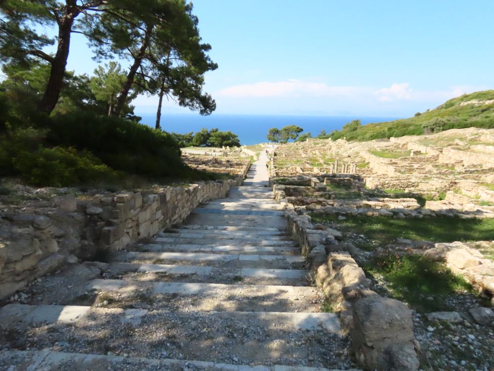 Looking down a path, partly stairs going down, with ruins on either side and the sea in the distance ahead.