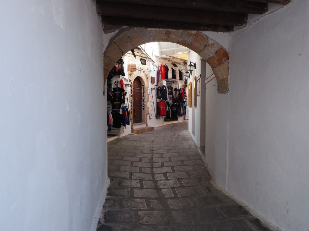 A narrow road with white walls on both sides, a stone arch and roof over part of it, shops visible ahead.