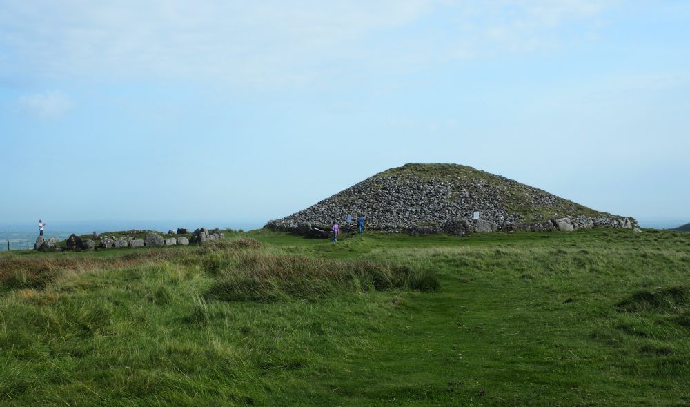 A large mound covered with rock and a smaller stone circle to the left of it. A big view in the background over the countryside below Loughcrew.