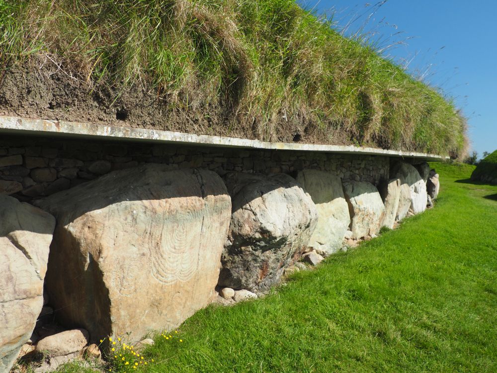 A view along the side of the tomb: stones face the bottom of the mound, grass covers everything about the stones. One stone has a carved spiral on its face.