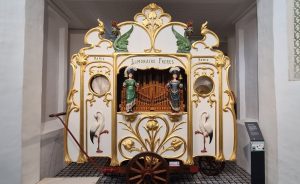 A street organ with two female figures in front of organ pipes and lots of painted decoration, much of it gold.