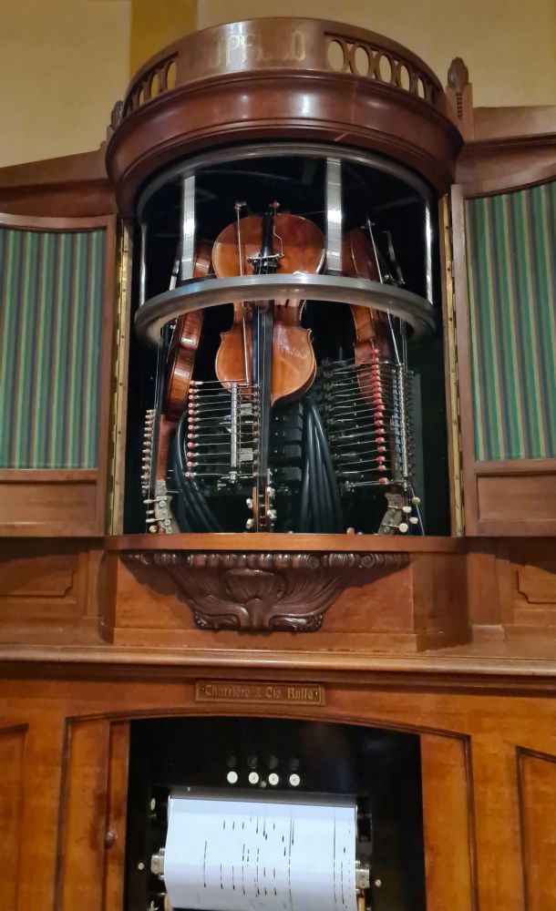 Below, the punched paper that rolls through the violina; above, the upright violins and circular bow, flanked by organ pipes.