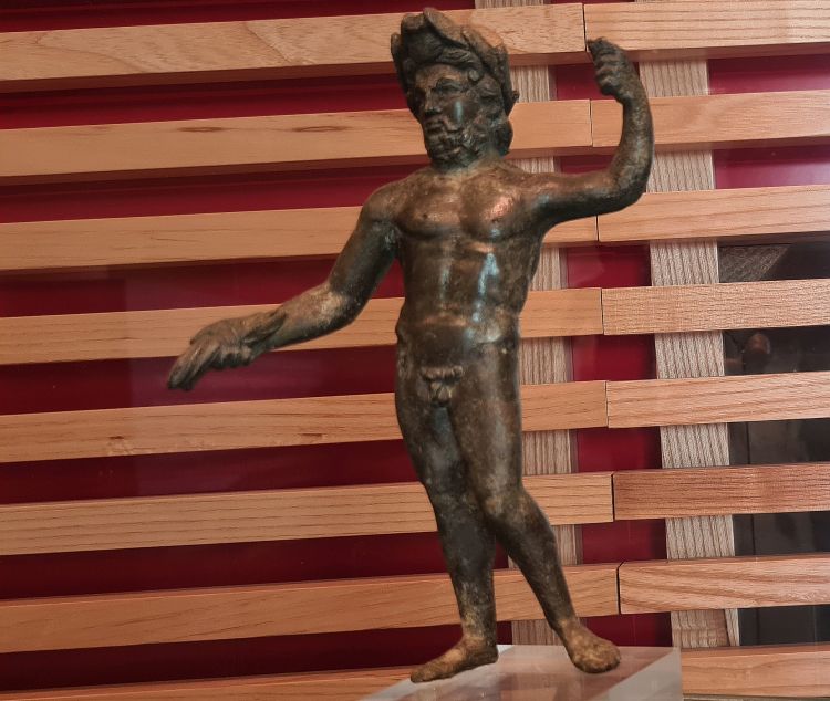 A small figurine of a naked man or god. One hand is outstretched in front of him; the other is raised.
