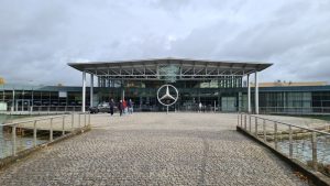 A modern building with a roof extending out from the front and a huge Mercedes-Benz logo standing in front of it in the center.