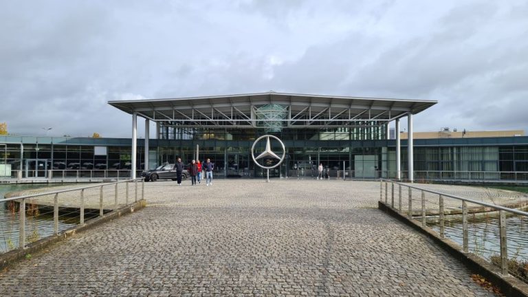 A modern building with a roof extending out from the front and a huge Mercedes-Benz logo standing in front of it in the center.