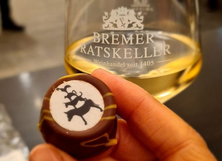 A glass of white wine with the words "Bremer Ratskeller." In front of it, a chocolate with an image of the Town Musicians of Bremen on it.