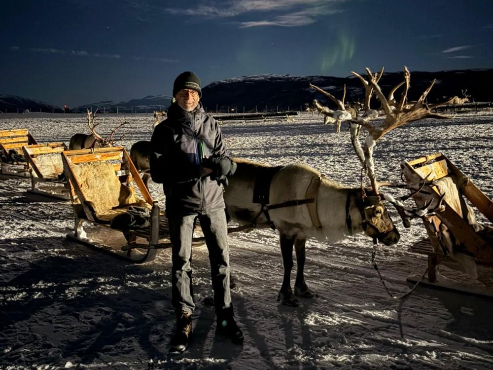 A man standing in front of a reindeer with huge antlers. The reindeer is harnessed to a sleigh.