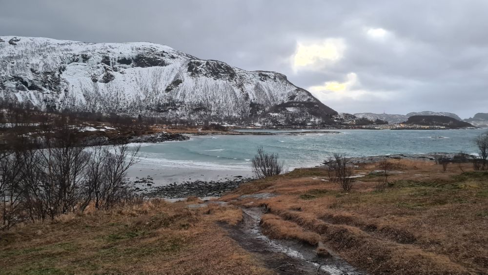 view over a half-moon beach, with a snow-covered hill beyond that. A grey, cloudy sky.