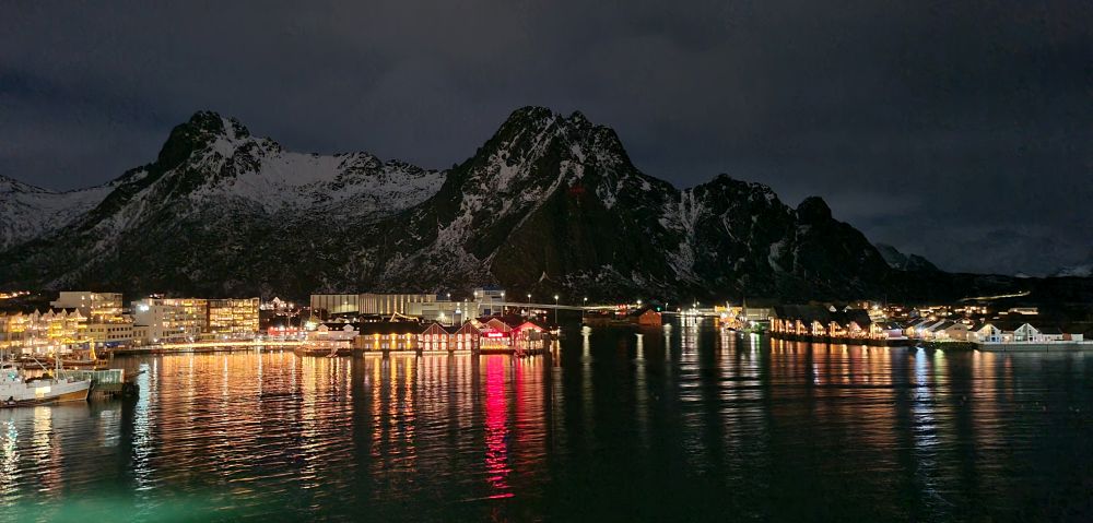 Nighttime shot of a small town, lots of lights from the buildings reflected in the water, with a mountain behind the town.