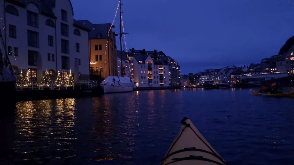 The tip of the kayak is visible and the dark water: dark houses lit with strings of light on either side.