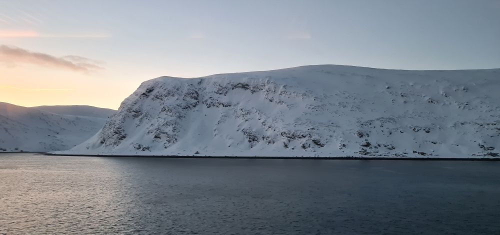 A snowy hill with steep sides rising straight from the sea, which is very smooth. Sunrise colors behind it.