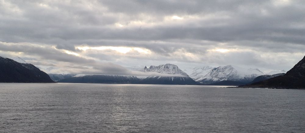 A large expanse of smooth sea, with mountains on the horizon topped with snow, clouds wrapped around their tops.