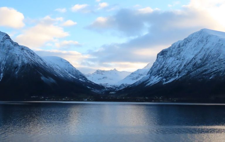 A view across the water at snow-covered mountains, with a small town nestled near the water between them.