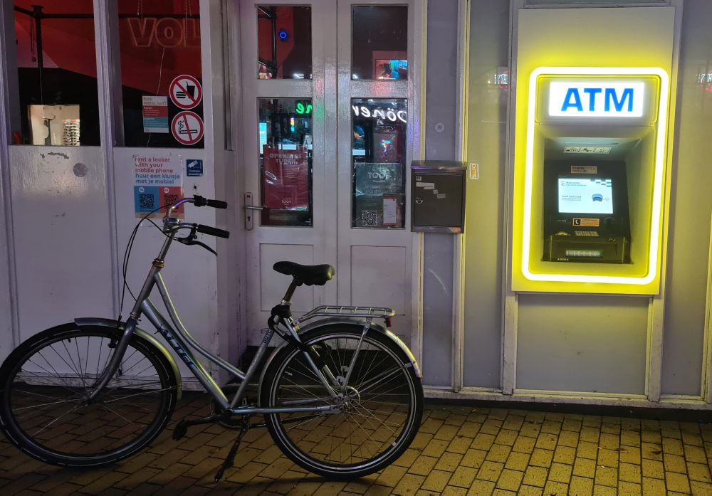 An ATM, brightly lit at night, with a bike parked next to it.