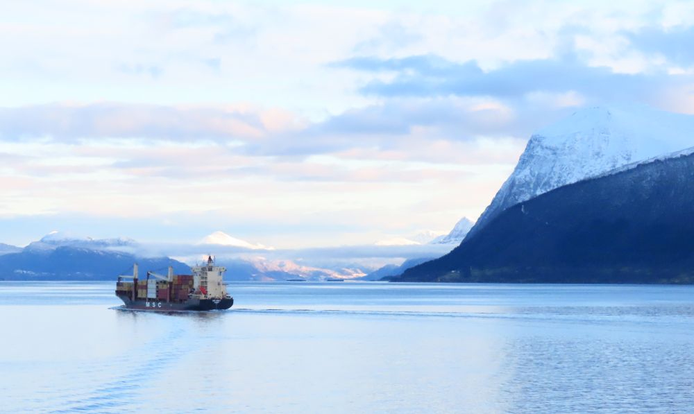 A smallish cargo ship on a calm sea, with snow-covered mountains in the background.