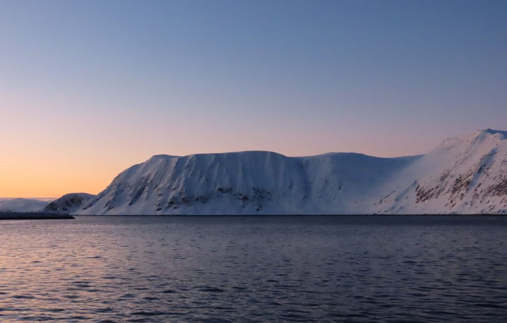 An entirely snow-covered flat-topped mountain with steep sides down to the water. Sunset sky.
