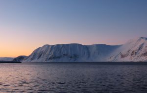 An entirely snow-covered flat-topped mountain with steep sides down to the water. Sunset sky.