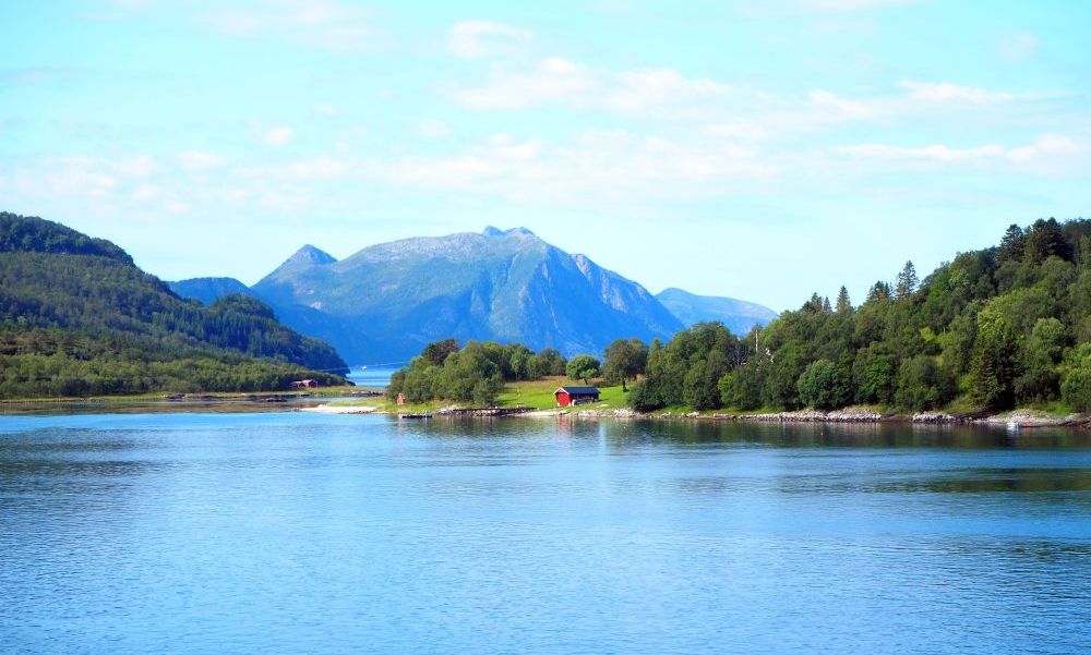 A red house, green field and trees on a point of land. Behind, a tall mountain with a few touches of snow.