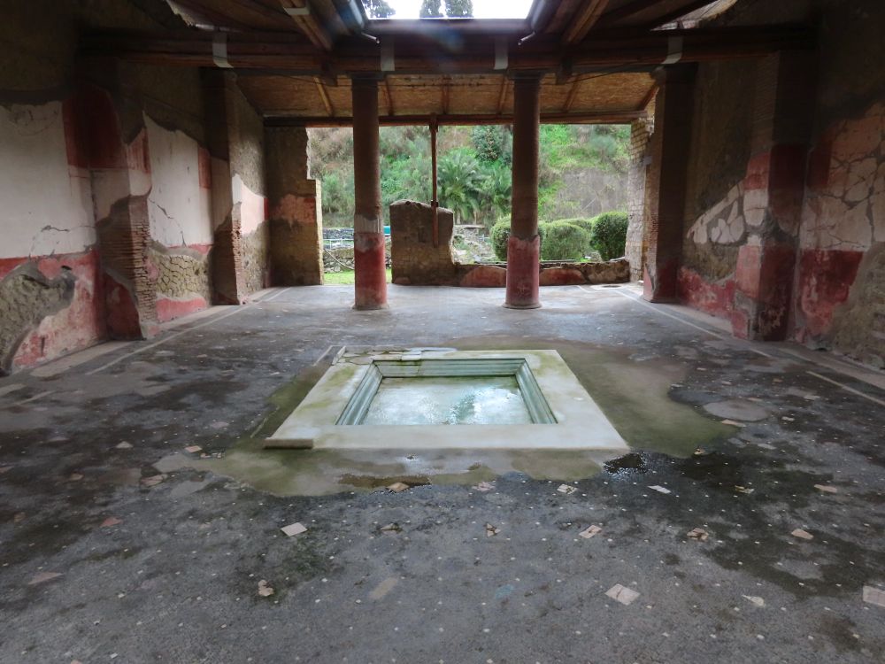 An open space with a shallow square pool in the center and pillars at the far end, which is open to the outdoors. The walls on the left and right have some remnants of frescoes.