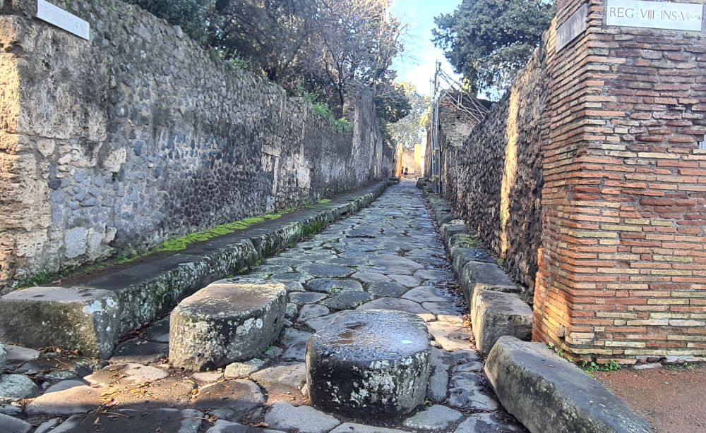 Looking down a narrow stone-paved street with narrow sidewalks on each side and stone walls lining the sidewalks. Two higher stones at the near end: an intersection.