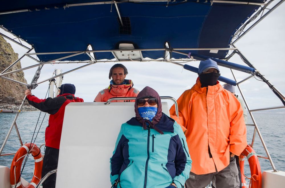 A participant sitting, bundled up, scientist beside her, also bundled up, as well as another person behind, all bundled up. In the center is the skipper.