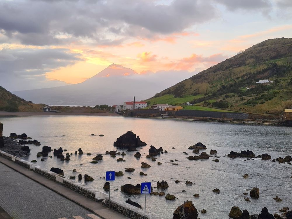 A bay with rock formations in it, a hill beyond with a white house, then a tall mountain in the distance behind that. Sunset colors.