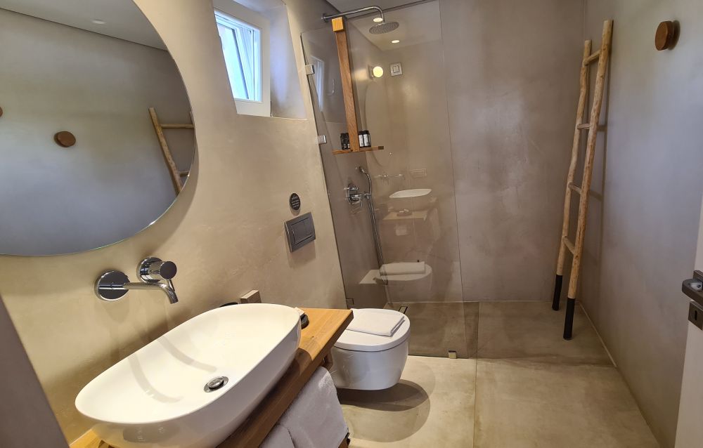 white sink on a wooden counter on the left, with a mirror above it. Toilet next to that, then a glass wall separating the shower space from the rest. All in sand-colored tile.