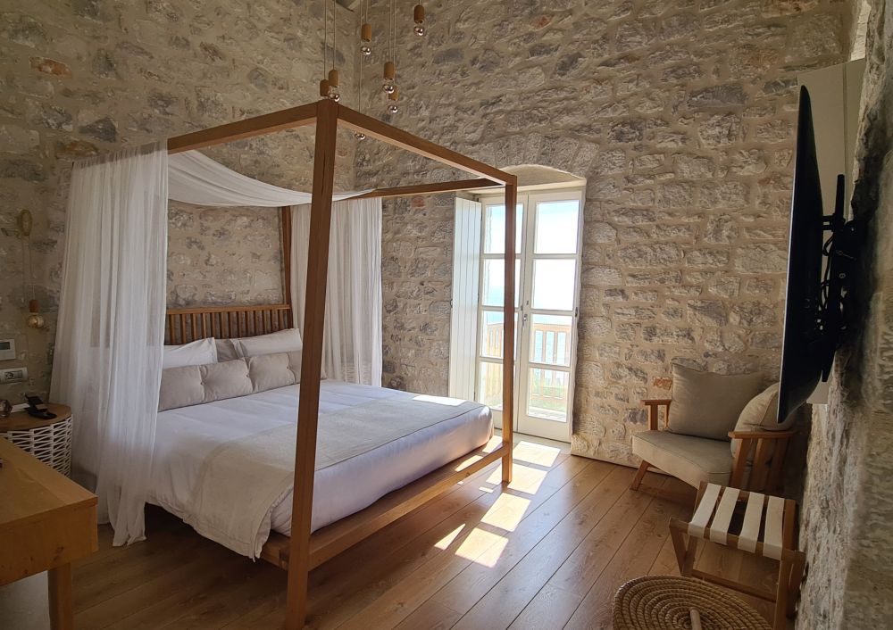 A large double canopy with white sheets and pillows and a filmy white cloth draped over as a canopy. Stone walls and a french door in the far wall letting in the light.