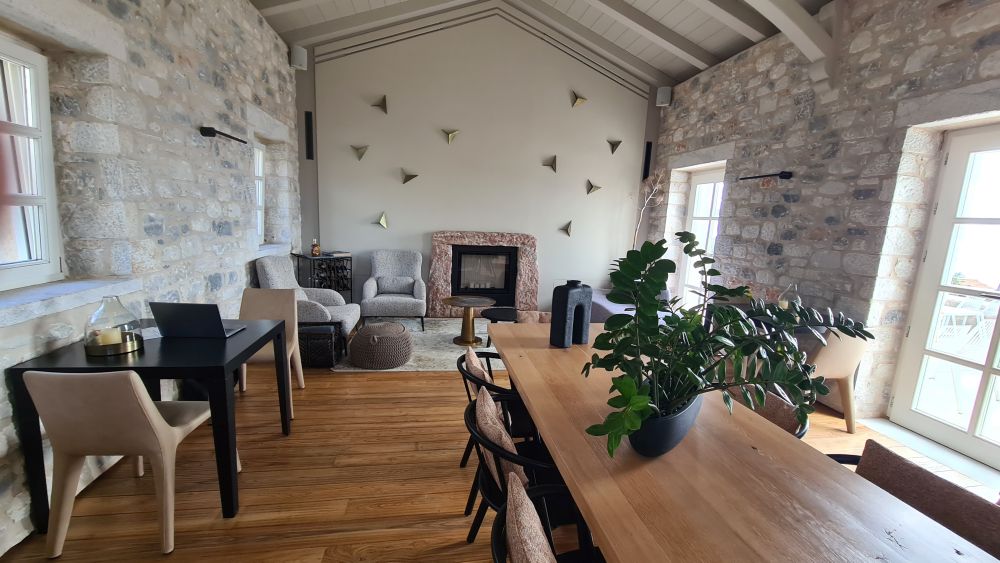 A long wooden table with chairs around it. Beyond that, a room with lots of light, a fireplace at the end of the room, and comfy chairs around it.