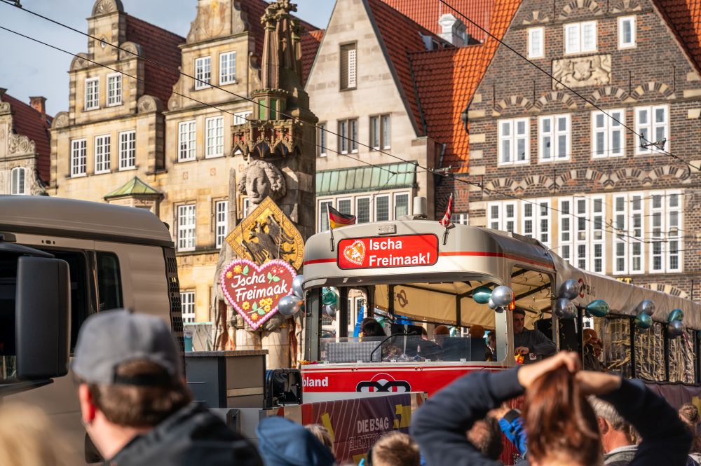 A decorated bus passing the Roland statue with crowds watching.