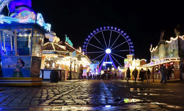 Looking down a cobbled street, brightly lit stalls on either side and a lit-up Ferris wheel in the background.