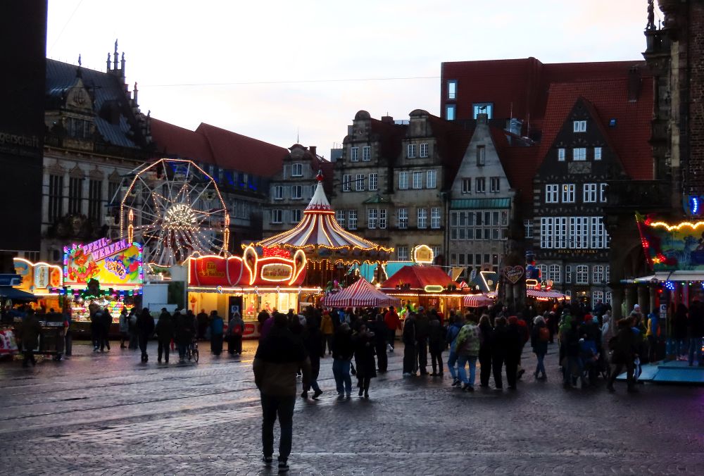 A square with old houses around it and brightly lit stalls filling it including a carousel and a small ferris wheel.