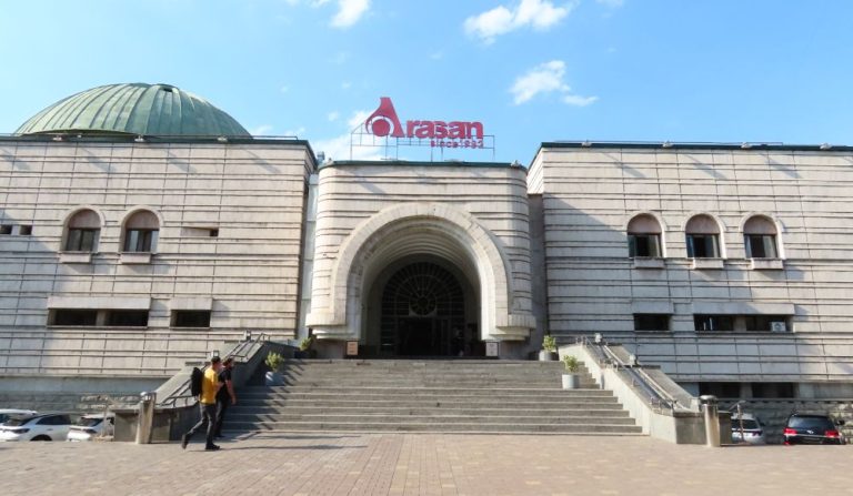 A white building with a wide stairway leading to its exit in an archway. The left-hand part has a dome.