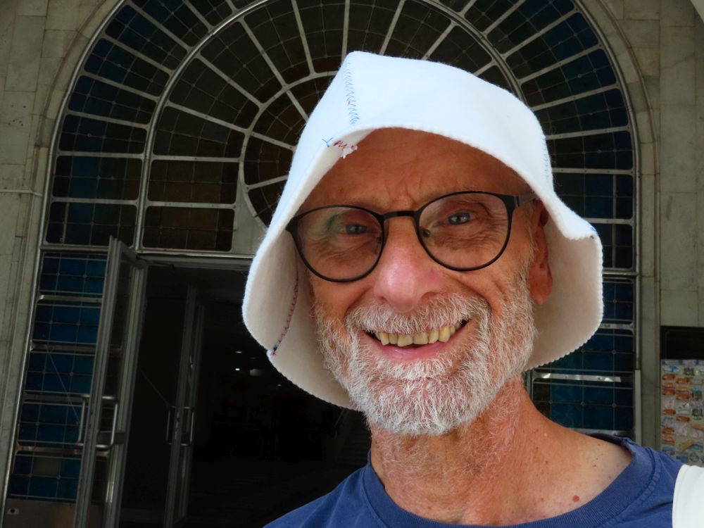 Smiling, gray beard and floppy white hat.