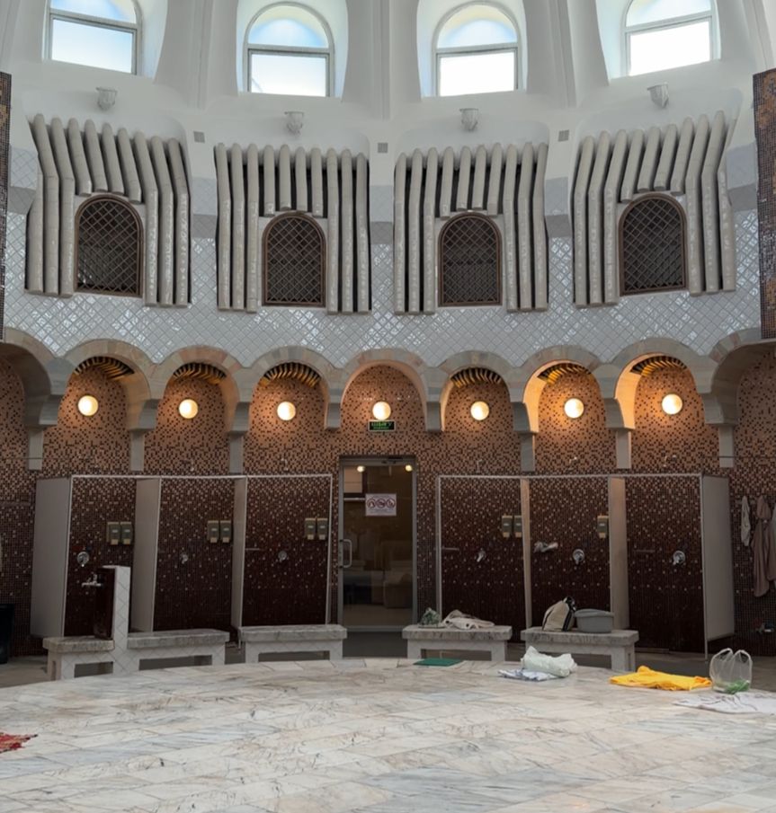 A curved wall with arches above a series of shower stalls with no doors. Above that, arched windows that are dark. Above them, another row of windows with the light shining through.