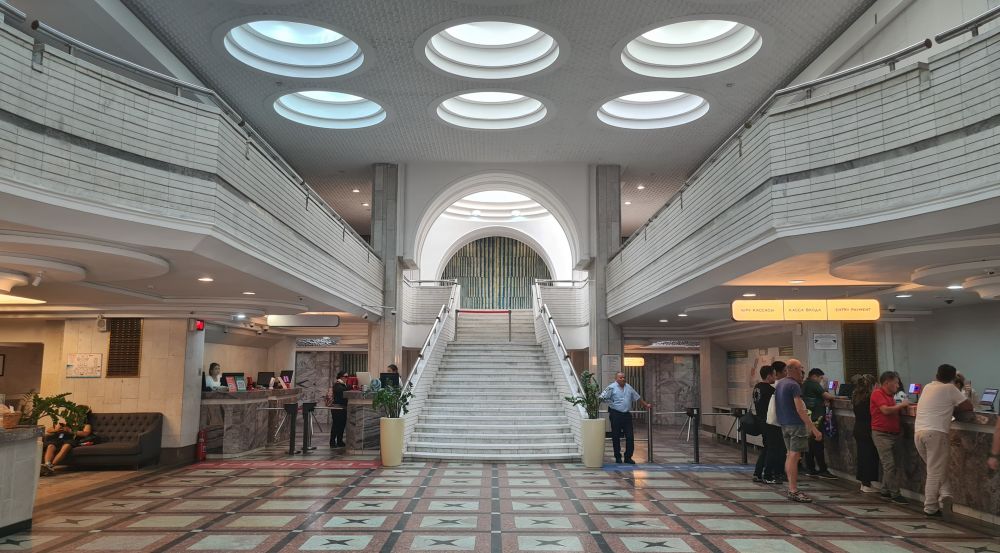 The lobby is lit by large round skylights in the ceiling. A grand stairway is straight ahead, and people cluster at the reception desk on the right.