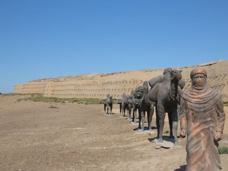 A life-sized sculpture of a man leading a caravan of camels. In the background, a long wall.