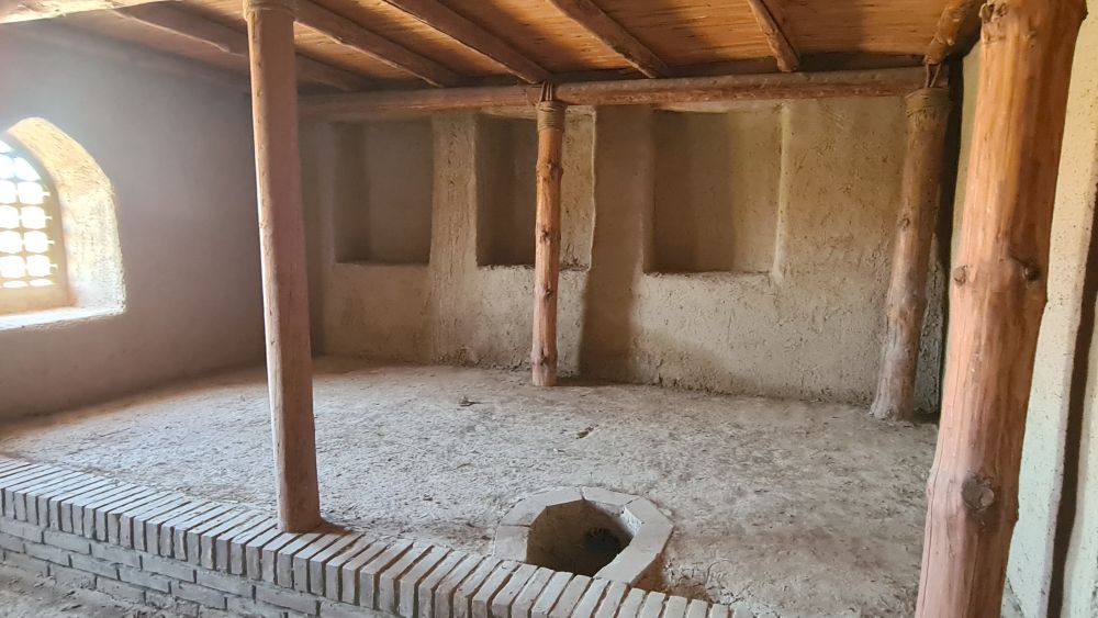 A room with mud walls and a wood-beamed ceiling. A small hole in the floor.
